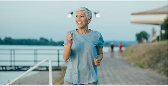A fit, older woman with short gray hair jogging along a waterfront path. She wears a light blue athletic shirt and smiles as she runs, with a scenic background featuring water, a railing, and other people in the distance A fit, older woman with short gray hair jogging along a waterfront path. She wears a light blue athletic shirt and smiles as she runs, with a scenic background featuring water, a railing, and other people in the distance
