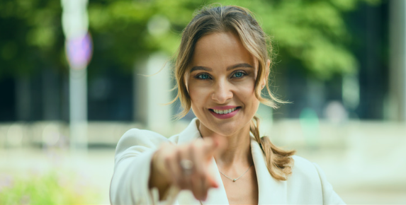 A smiling woman in a white blazer stands outdoors in a city park or urban environment. She is pointing directly at the camera with a confident and friendly expression, symbolizing encouragement, leadership, or motivation A smiling woman in a white blazer stands outdoors in a city park or urban environment. She is pointing directly at the camera with a confident and friendly expression, symbolizing encouragement, leadership, or motivation