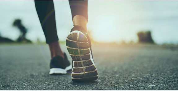 A close-up of a person’s feet wearing running shoes as they walk or jog on an outdoor road. The image is taken from a low angle, with the sun creating a lens flare effect, conveying movement, fitness, and determination A close-up of a person’s feet wearing running shoes as they walk or jog on an outdoor road. The image is taken from a low angle, with the sun creating a lens flare effect, conveying movement, fitness, and determination