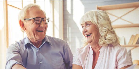 A smiling elderly couple sits together in a well-lit room, laughing and looking at each other with joy. The man wears glasses and a light blue shirt, while the woman has white hair and wears a pink blouse. The scene conveys happiness, love, and companionship in later life A smiling elderly couple sits together in a well-lit room, laughing and looking at each other with joy. The man wears glasses and a light blue shirt, while the woman has white hair and wears a pink blouse. The scene conveys happiness, love, and companionship in later life
