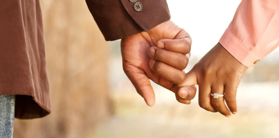 A close-up of a couple holding hands, with their fingers gently intertwined. The woman wears an engagement ring, suggesting themes of love, commitment, or marriage A close-up of a couple holding hands, with their fingers gently intertwined. The woman wears an engagement ring, suggesting themes of love, commitment, or marriage