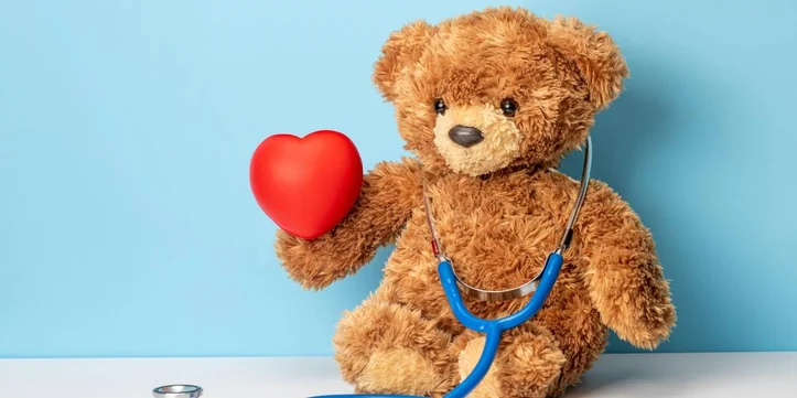 A fluffy brown teddy bear is sitting against a bright blue background, wearing a stethoscope around its neck and holding a red heart in one paw. The scene gives the impression of a doctor teddy bear promoting health or care. A fluffy brown teddy bear is sitting against a bright blue background, wearing a stethoscope around its neck and holding a red heart in one paw. The scene gives the impression of a doctor teddy bear promoting health or care.