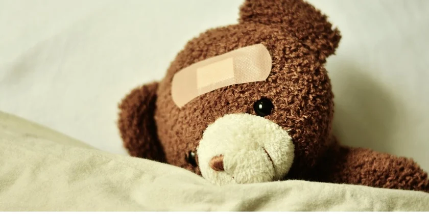 A brown teddy bear with a beige bandage on its forehead is lying down on a soft surface, partially covered by a blanket. The bear's black eyes and slightly tilted head give it a sad or injured appearance. A brown teddy bear with a beige bandage on its forehead is lying down on a soft surface, partially covered by a blanket. The bear's black eyes and slightly tilted head give it a sad or injured appearance.