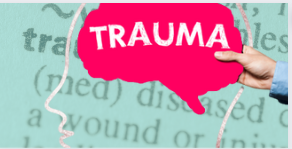A visual representation of trauma and mental health. The image features a pink speech bubble with the word "TRAUMA" written in bold white letters, held by a hand. The background consists of a faded dictionary-style definition of the word "trauma" along with an outlined silhouette of a human head. A visual representation of trauma and mental health. The image features a pink speech bubble with the word "TRAUMA" written in bold white letters, held by a hand. The background consists of a faded dictionary-style definition of the word "trauma" along with an outlined silhouette of a human head.