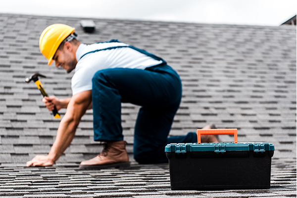 Roofer in a hard hat repairing shingles with a toolbox nearby, highlighting the comparison between DIY and professional roof replacement. Roofer in a hard hat repairing shingles with a toolbox nearby, highlighting the comparison between DIY and professional roof replacement.