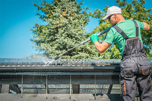 Worker using a pressure washer to clean debris from gutters on a roof, ensuring proper maintenance and preventing water damage. Worker using a pressure washer to clean debris from gutters on a roof, ensuring proper maintenance and preventing water damage.