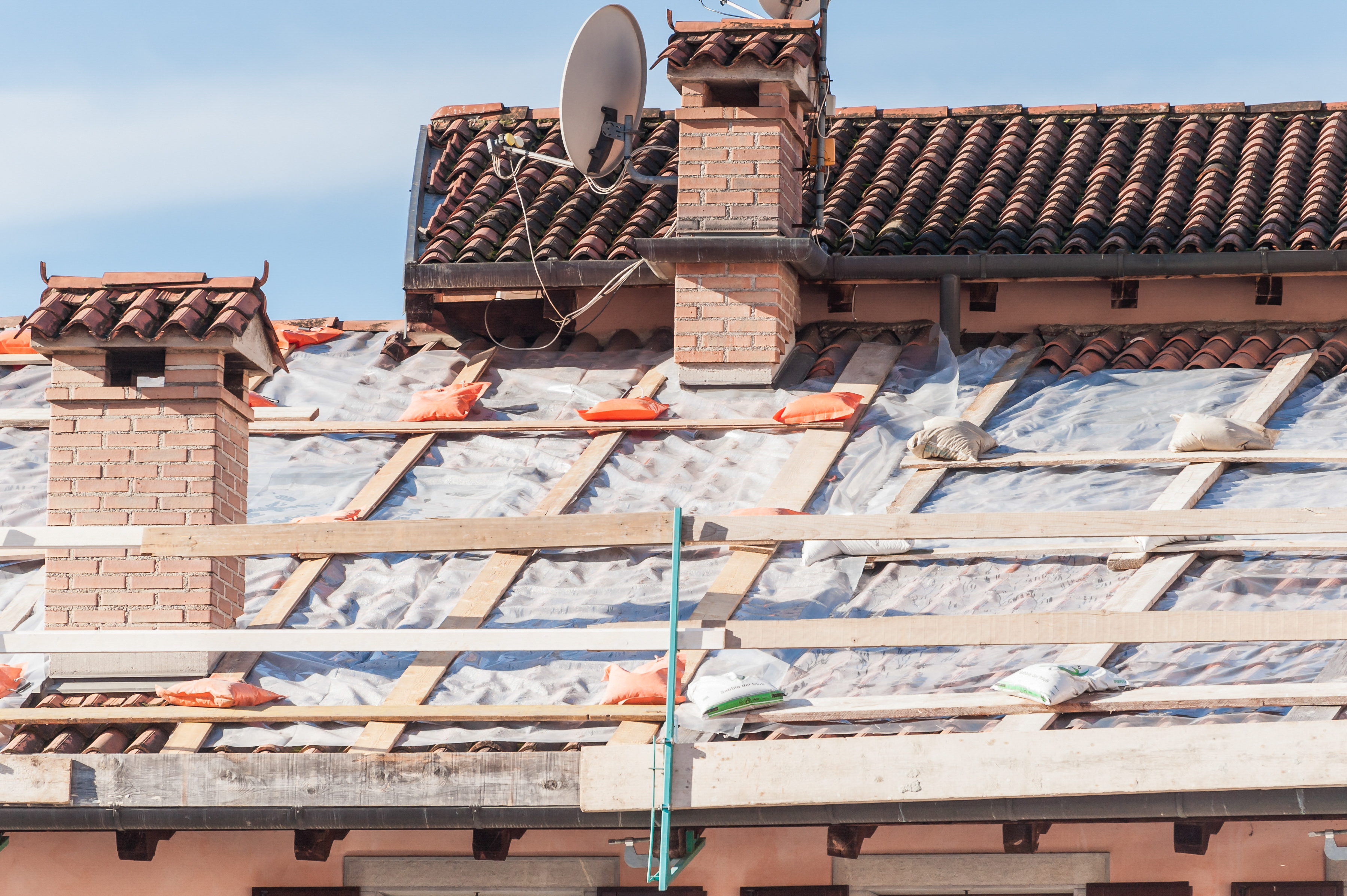 Roof under repair with exposed wooden framework, plastic sheeting, and sandbags, highlighting roof damage and restoration process. Roof under repair with exposed wooden framework, plastic sheeting, and sandbags, highlighting roof damage and restoration process.
