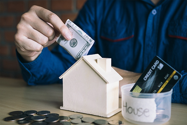 Close-up of a person inserting cash into a miniature wooden house model, symbolizing home investment, budgeting, or real estate savings. Close-up of a person inserting cash into a miniature wooden house model, symbolizing home investment, budgeting, or real estate savings.