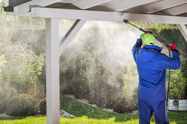 A professional cleaner wearing a blue waterproof suit and green cap uses a high-pressure washer to clean an outdoor patio roof, removing dirt, debris, and mildew. A professional cleaner wearing a blue waterproof suit and green cap uses a high-pressure washer to clean an outdoor patio roof, removing dirt, debris, and mildew.