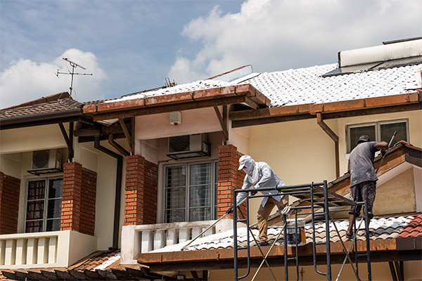 Two workers wearing protective gear are standing on scaffolding, repairing or painting the white-tiled roof of a residential building with red brick accents. The house has multiple balconies and air conditioning units mounted on the walls. The workers are engaged in maintenance or restoration work to improve the roof’s durability and appearance. Two workers wearing protective gear are standing on scaffolding, repairing or painting the white-tiled roof of a residential building with red brick accents. The house has multiple balconies and air conditioning units mounted on the walls. The workers are engaged in maintenance or restoration work to improve the roof’s durability and appearance.