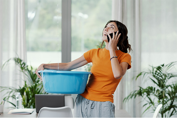 A woman holding a blue bucket to catch water from a leaking ceiling while making a phone call, highlighting urgent roofing issues. A woman holding a blue bucket to catch water from a leaking ceiling while making a phone call, highlighting urgent roofing issues.