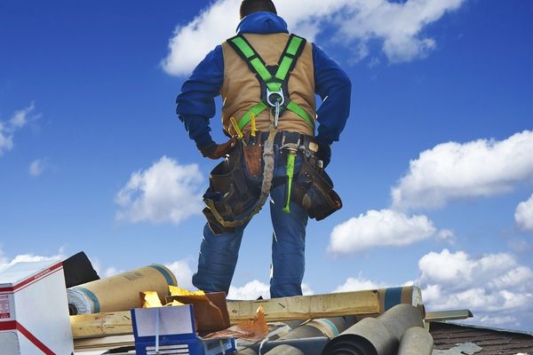 A professional roofer standing on a rooftop with safety gear, surrounded by roofing materials, under a bright blue sky. A professional roofer standing on a rooftop with safety gear, surrounded by roofing materials, under a bright blue sky.