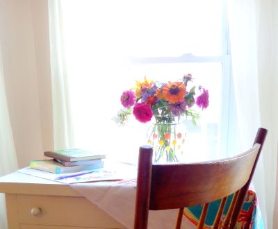 An image of sunlight filtering through a sheer curtain onto a small wooden writing desk with books, paper, calligraphy pen, and a vase of fresh flowers, and a wooden chair.