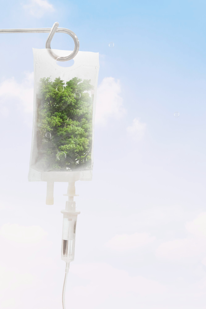An intravenous (IV) bag hangs from a metal hook against a bright blue sky with scattered clouds. Instead of containing fluid, the IV bag holds a small, lush green tree or plant, and medical tubing extends from the bottom. The image combines clinical and natural elements, symbolizing the fusion of wellness, nature, and healthcare.