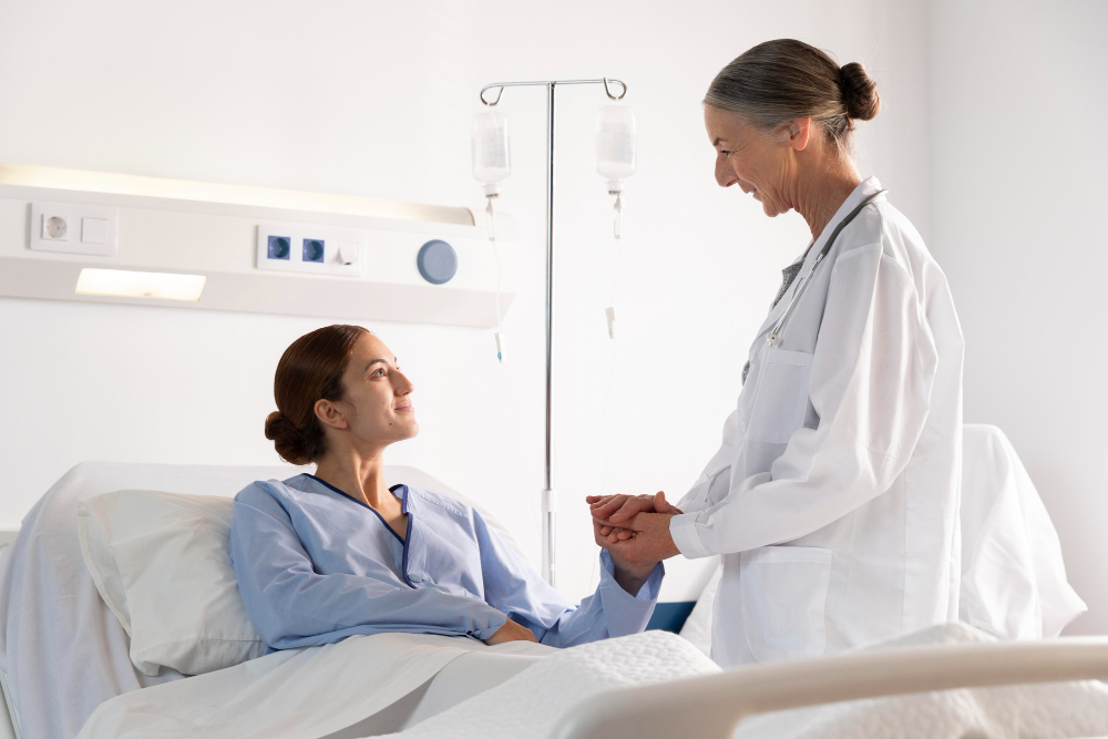 Hospital room scene featuring a patient lying in bed wearing a light blue gown, with a healthcare professional in a white lab coat gently holding the patient’s hand. An IV stand with two hanging IV bags stands beside the bed.