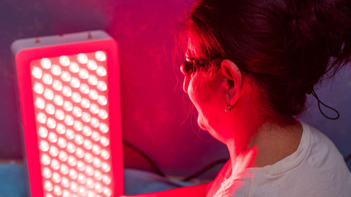 Woman sitting in front of a vertical red light therapy panel, wearing protective goggles and receiving treatment on her face and upper body in a home or clinic setting. Woman sitting in front of a vertical red light therapy panel, wearing protective goggles and receiving treatment on her face and upper body in a home or clinic setting.