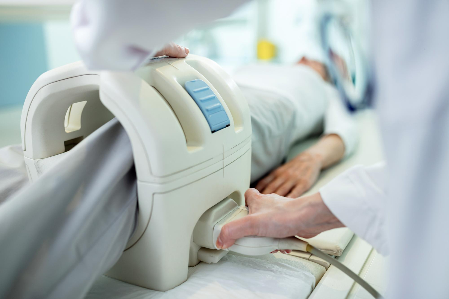 A patient lies on a medical imaging table while a healthcare professional adjusts a large cylindrical scanning device, likely an MRI or CT machine, in a clinical setting. A patient lies on a medical imaging table while a healthcare professional adjusts a large cylindrical scanning device, likely an MRI or CT machine, in a clinical setting.