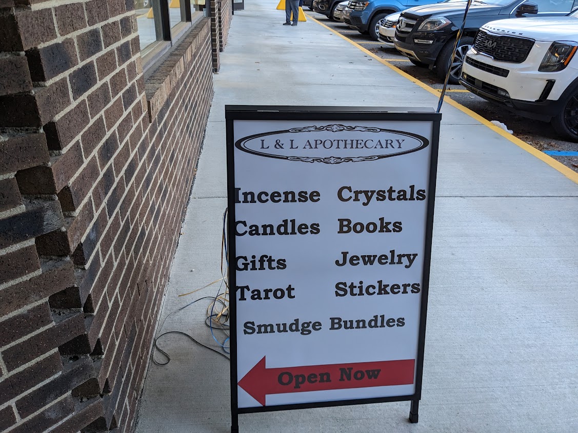a sandwich board sign on the sidewalk, with L&L Apothecary at the top and a list of items they sell: incense, crystals, candles, books, gifts, jewelry, tarot, stickers, smudge bundles