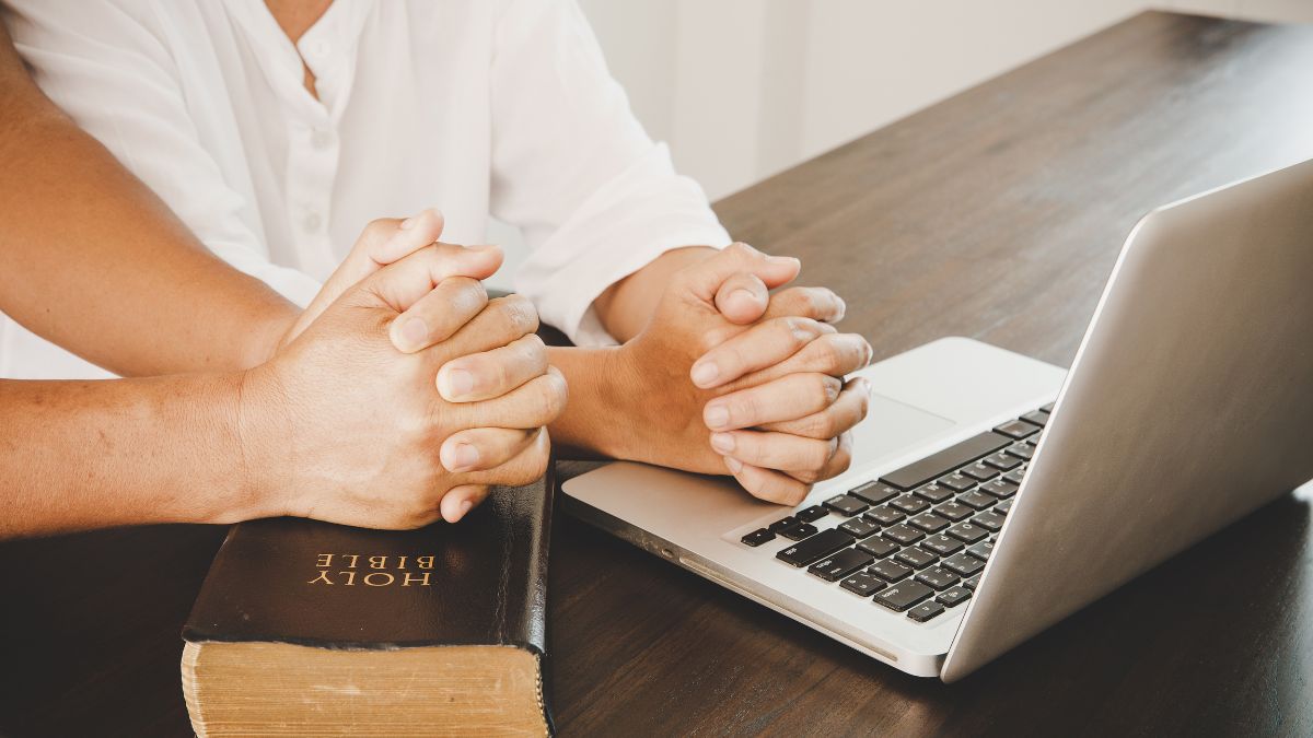 Person resting their clasped hands on laptop and another resting their hands on a Bible as concept for virtual worship.