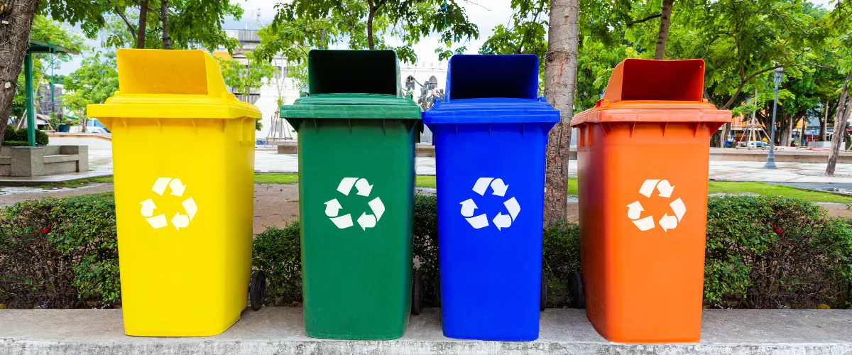 Four color-coded recycling bins — yellow, green, blue, and orange — in a public park setting