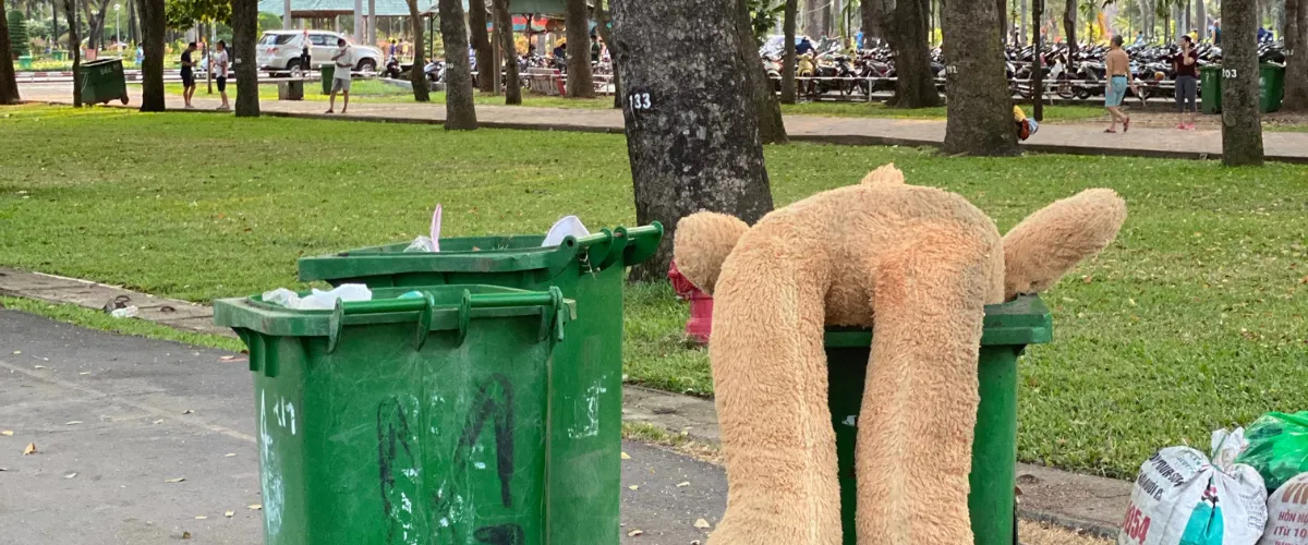 Green trash bins in a park area with a large plush teddy bear discarded across the bin. Green trash bins in a park area with a large plush teddy bear discarded across the bin.