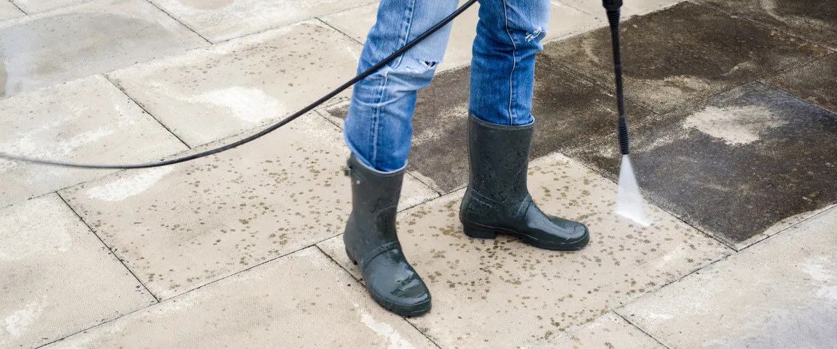 Worker wearing rubber boots pressure washing dirty outdoor pavers.