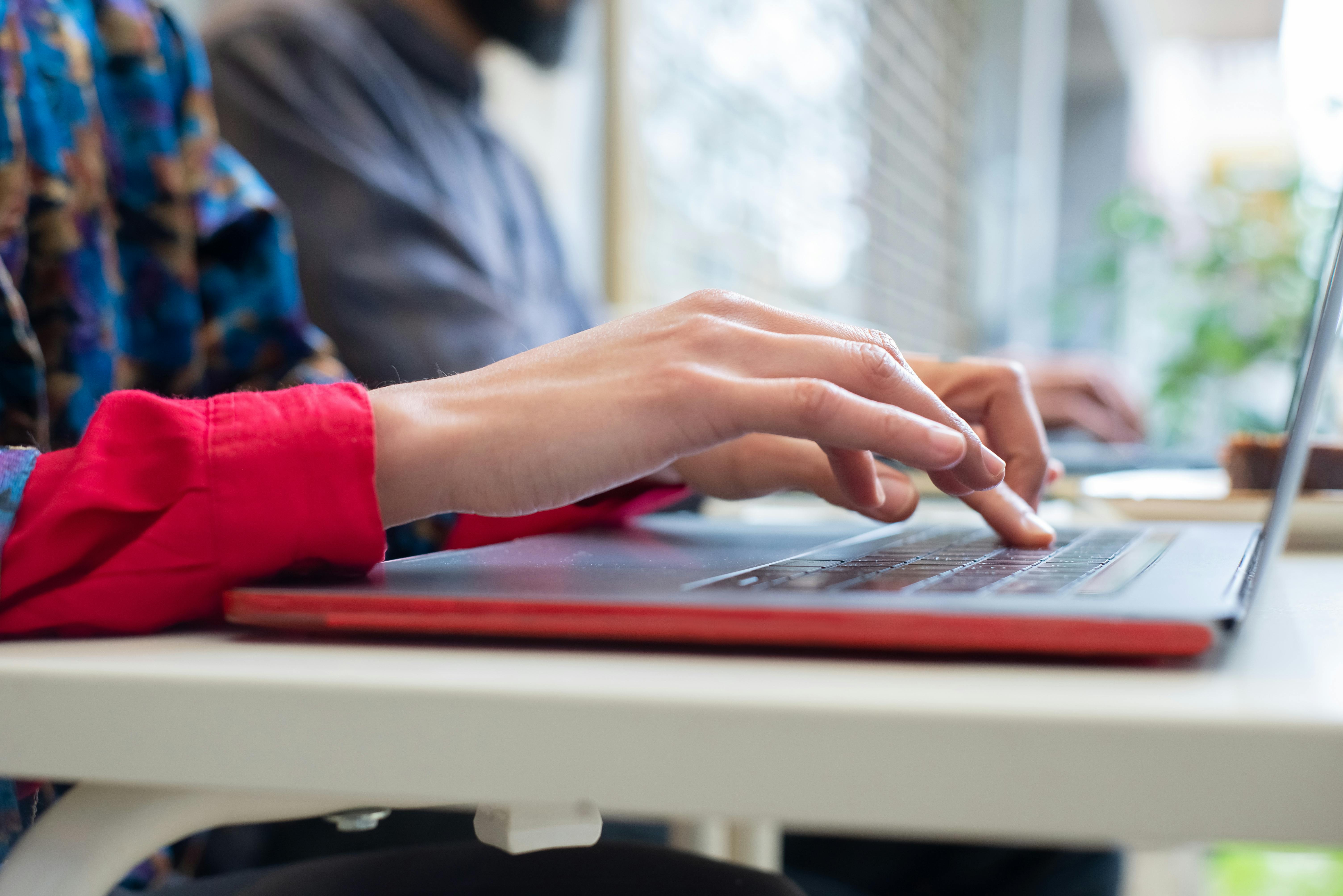 Close-up of a person typing on a laptop with a red case, working at an outdoor table with another individual in the background. Close-up of a person typing on a laptop with a red case, working at an outdoor table with another individual in the background.