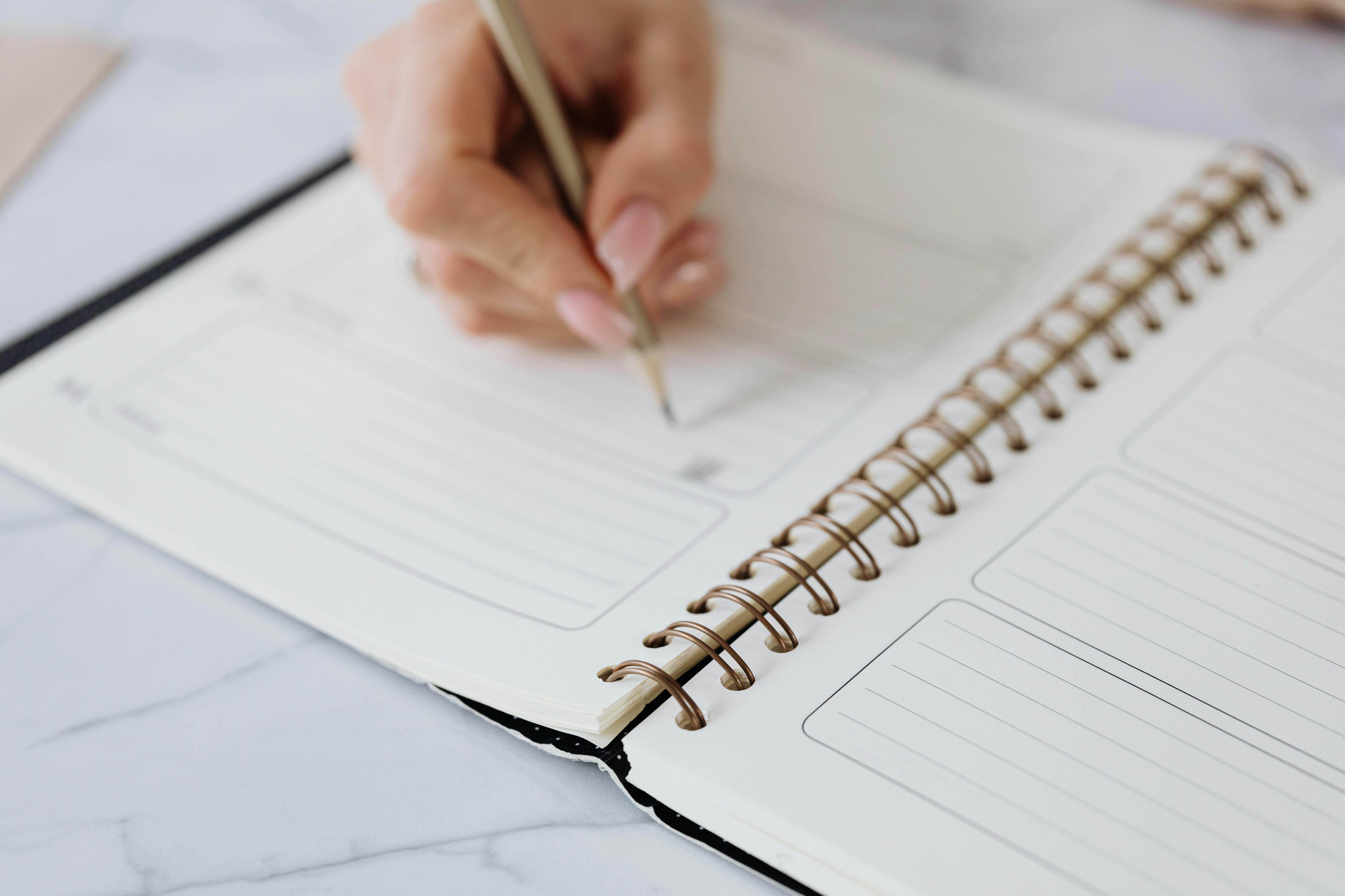 Close-up of a person writing in a spiral-bound planner with a gold pen, filling out lined sections on a marble surface. Close-up of a person writing in a spiral-bound planner with a gold pen, filling out lined sections on a marble surface.