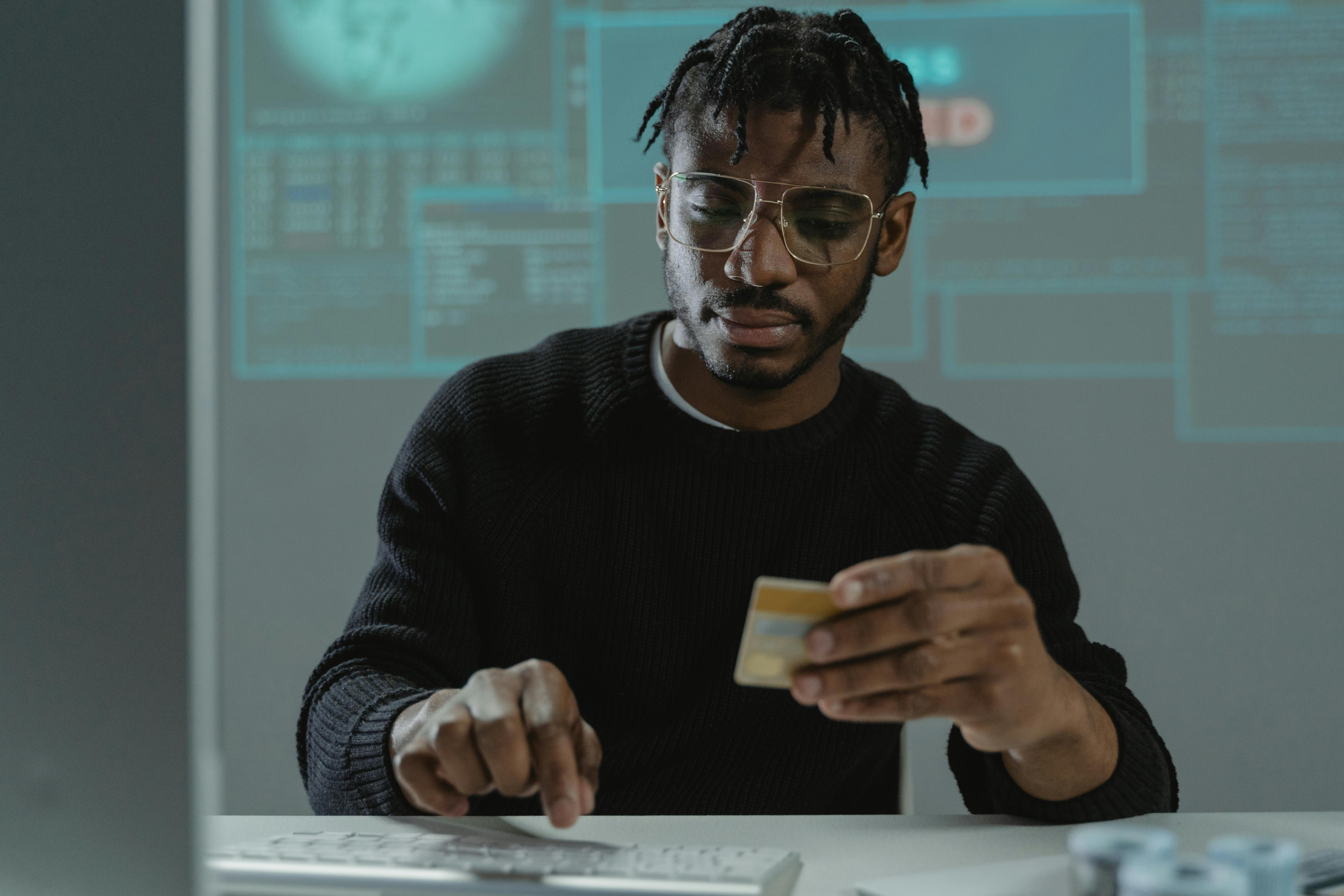 Man with glasses and a black sweater using a credit card for an online transaction, with cybersecurity data on a screen behind him. Man with glasses and a black sweater using a credit card for an online transaction, with cybersecurity data on a screen behind him.