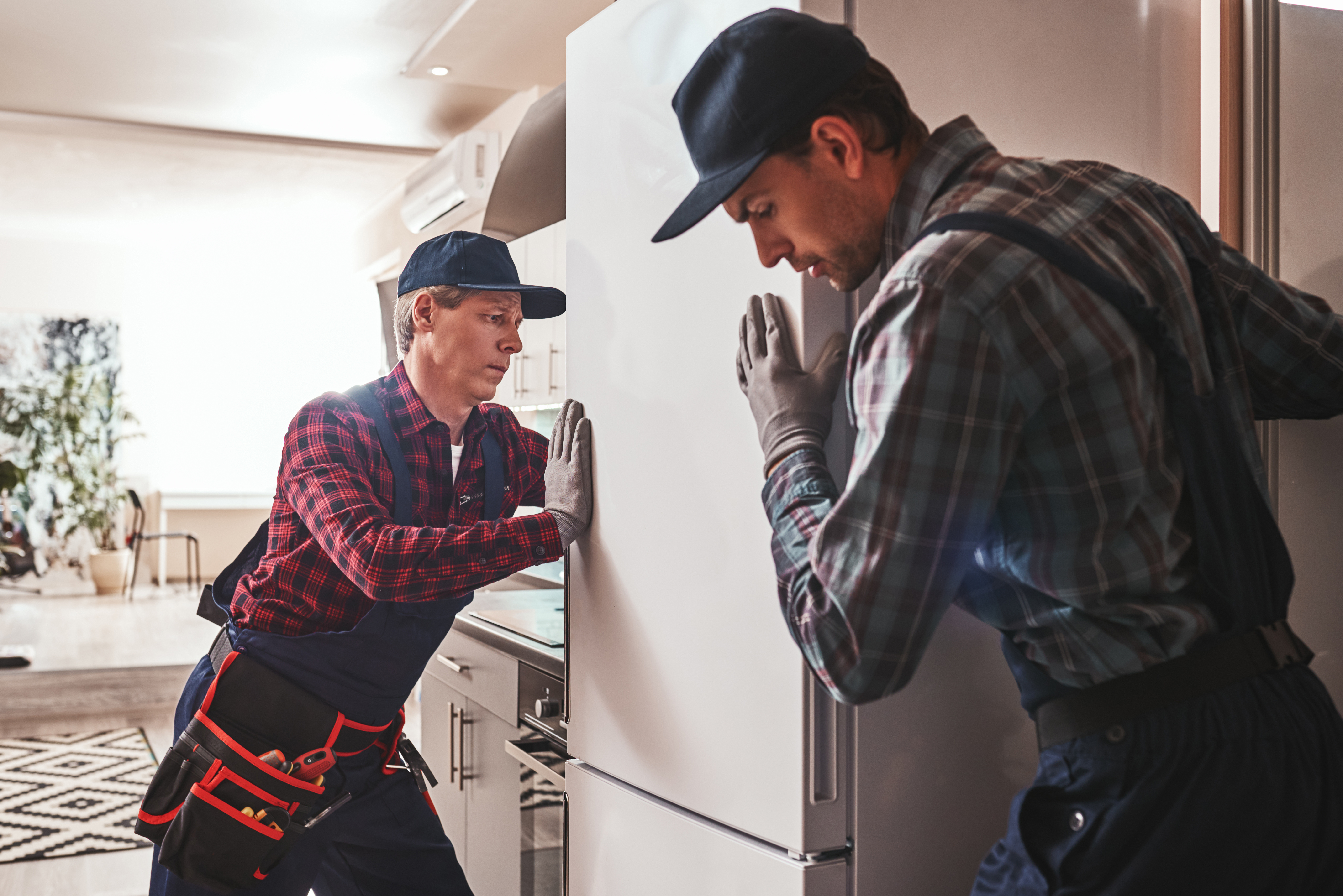 Two men in overalls and hard hats repairing a refrigerator.