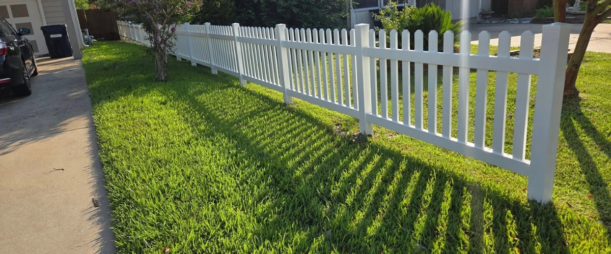 Newly installed white picket fence along a backyard lawn casting long shadows. Newly installed white picket fence along a backyard lawn casting long shadows.