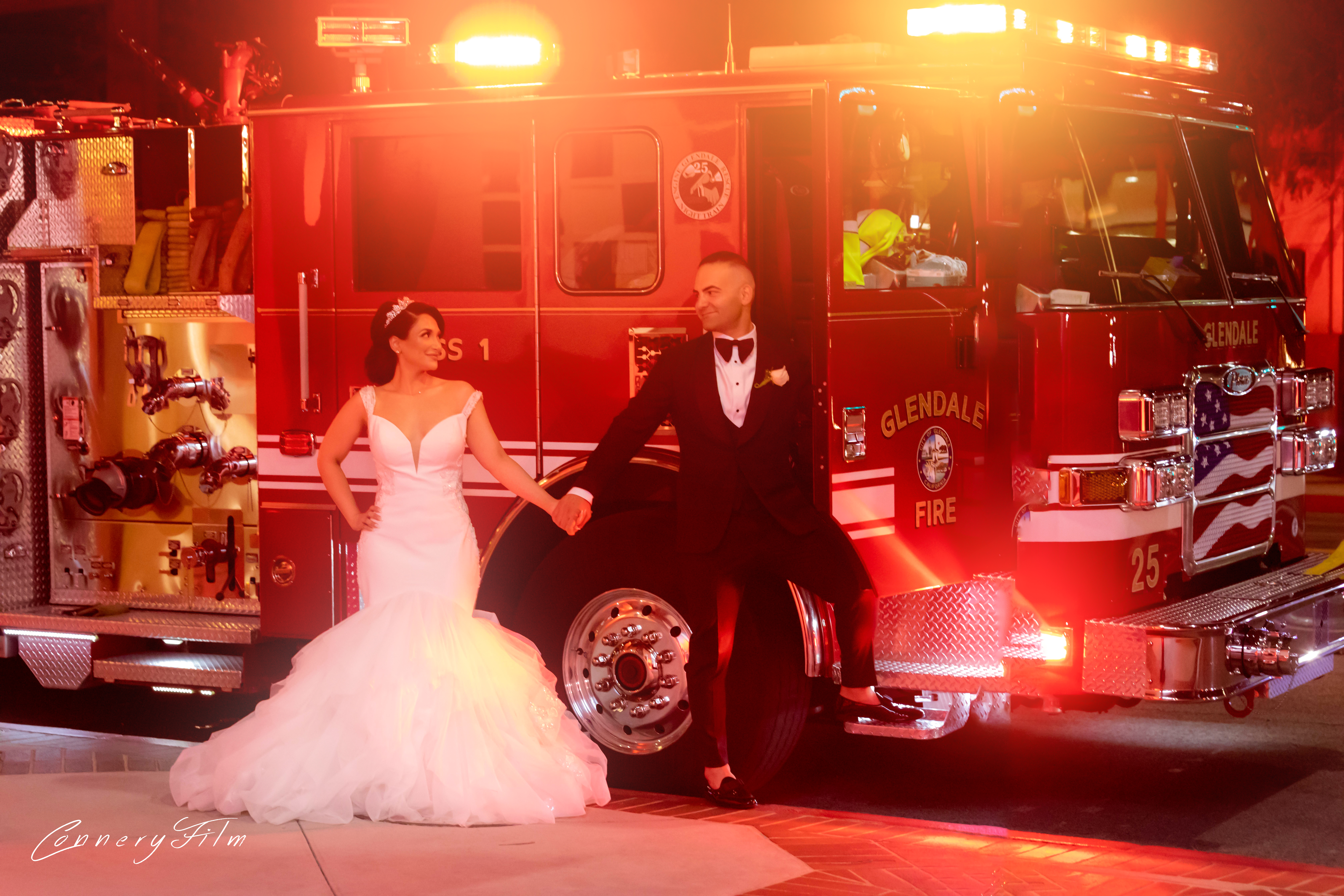 A bride in a white mermaid gown and a groom in a tuxedo holding hands and posing playfully in front of a red Glendale Fire Department truck at night, with bright lights.