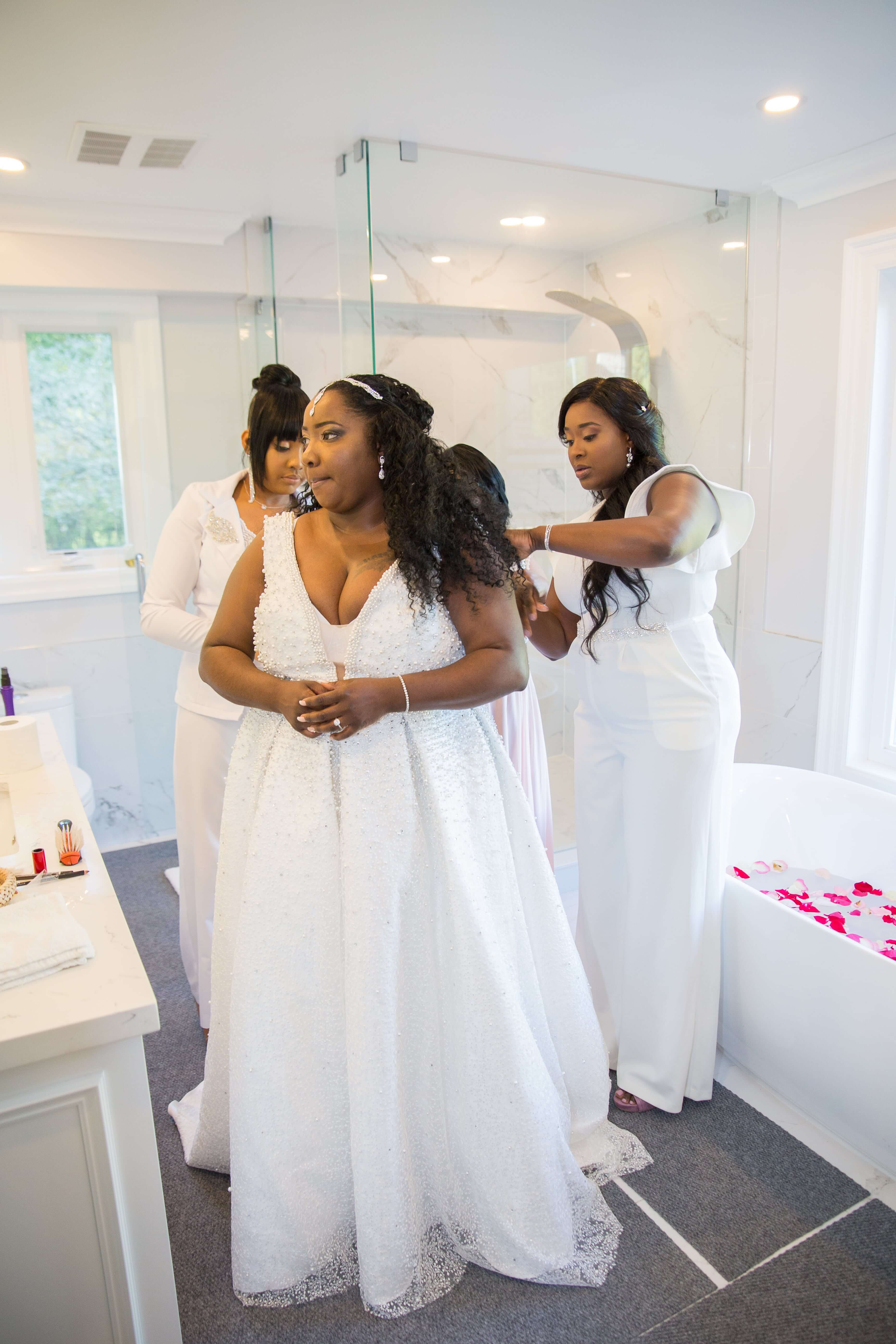 Black women getting ready in bathroom with friends helping her put on wedding dress