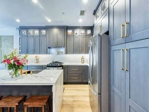 A stylish kitchen with painting gray-blue cabinets, a marble island with wooden stools, and a vase of flowers under bright lighting.
