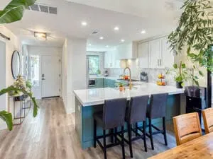 A modern kitchen with painting teal cabinets, a white island with black stools, and lush indoor plants under bright lighting.