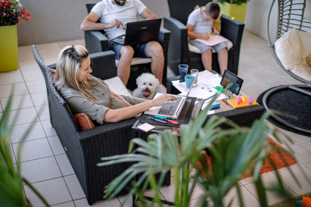 A diverse group of people, different ages and races, working remotely at home on laptops in a living room or home office, with a cute small dog lying next to one of them, with headphones on their ears.