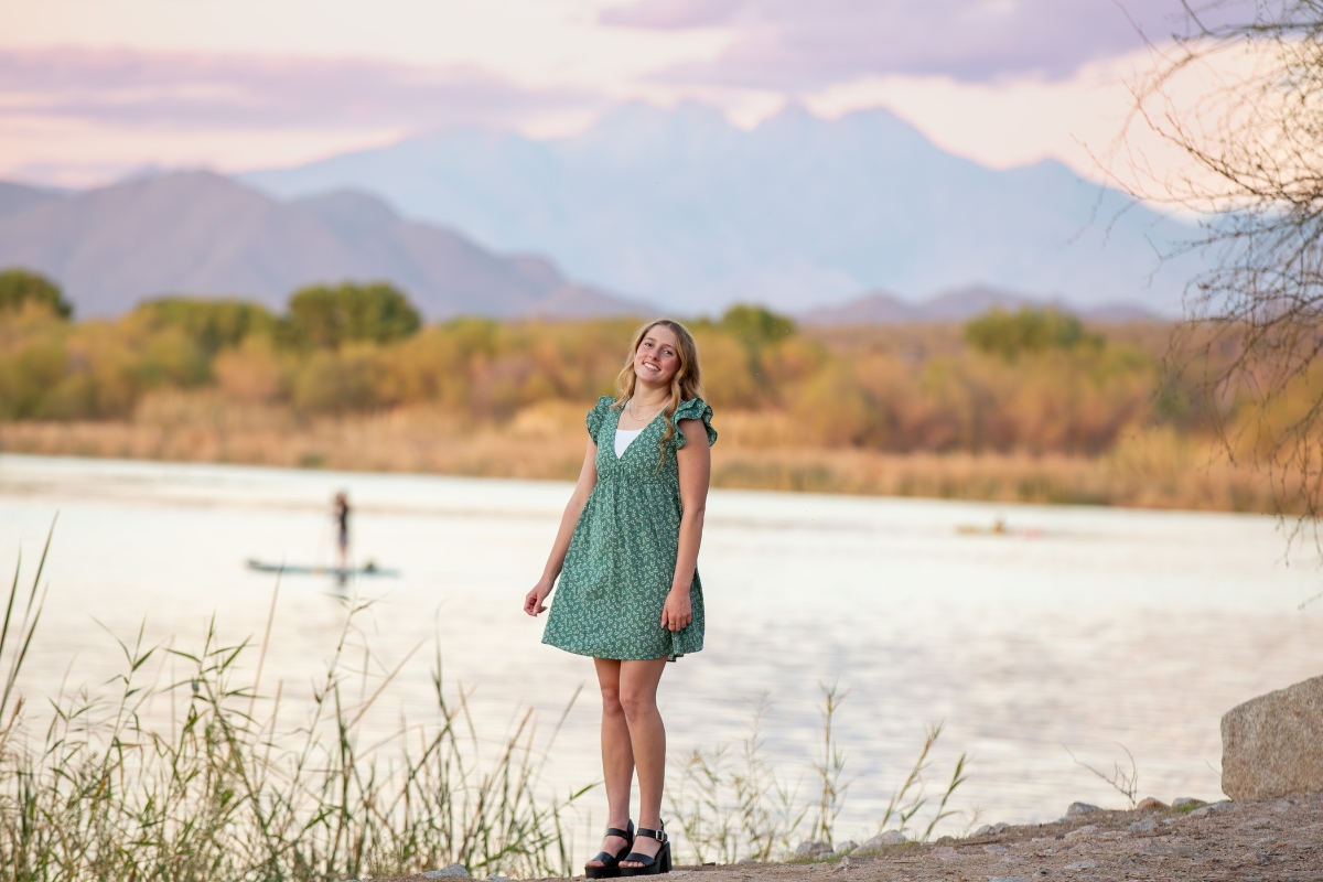 Senior girl in green dress standing by the water at Coon Bluff in Mesa, Arizona, photographed by Gilbert senior photographer Andria May Photography, with mountains and paddleboarders in the background. Senior girl in green dress standing by the water at Coon Bluff in Mesa, Arizona, photographed by Gilbert senior photographer Andria May Photography, with mountains and paddleboarders in the background.