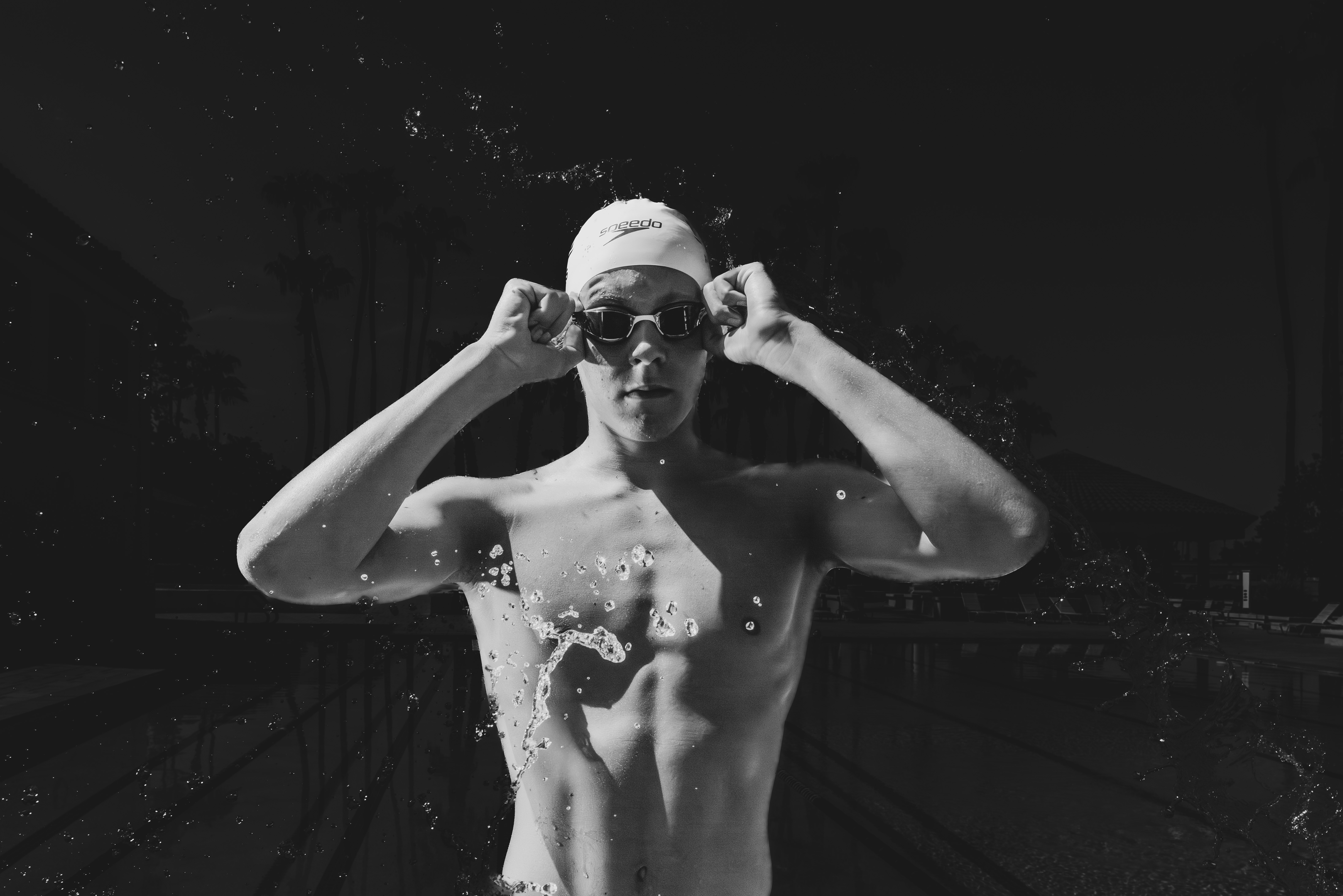 Dramatic black-and-white senior sports portrait of a male swimmer adjusting his goggles as water splashes around him, photographed at an outdoor pool in Gilbert, Arizona by Gilbert senior photographer Andria May Photography.