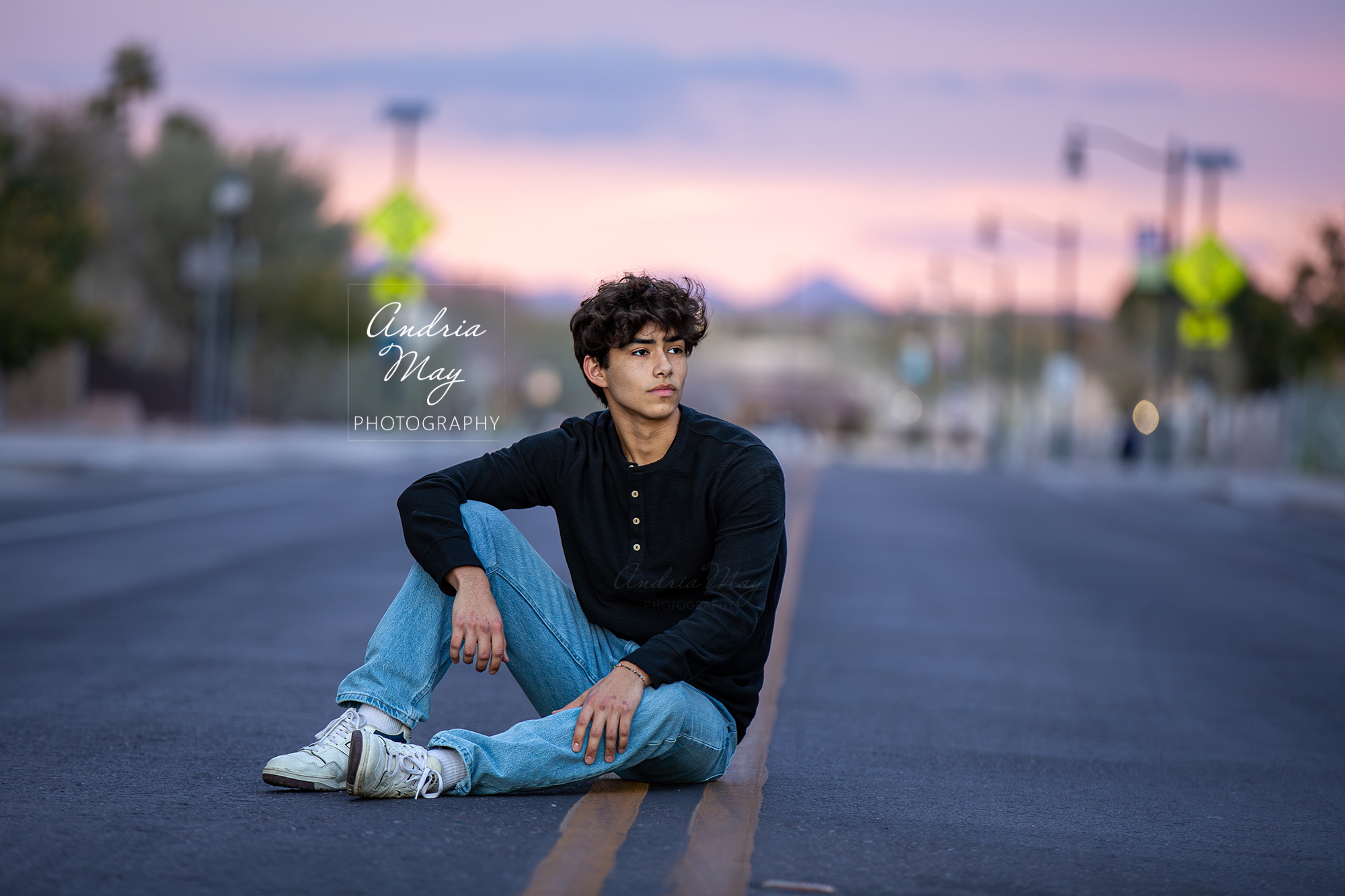 Senior portrait of a high school boy sitting in the street in Downtown Gilbert at sunset, wearing jeans and a black shirt. The vivid Arizona sky creates a dramatic backdrop, photographed by Andria May Photography. Senior portrait of a high school boy sitting in the street in Downtown Gilbert at sunset, wearing jeans and a black shirt. The vivid Arizona sky creates a dramatic backdrop, photographed by Andria May Photography.