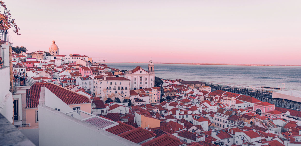 Uma vista aérea, ao final da tarde, de uma zona nobre e moderna de Lisboa ou do Porto, onde se veem edifícios residenciais de luxo junto a uma marina ou a um parque verde bem cuidado.