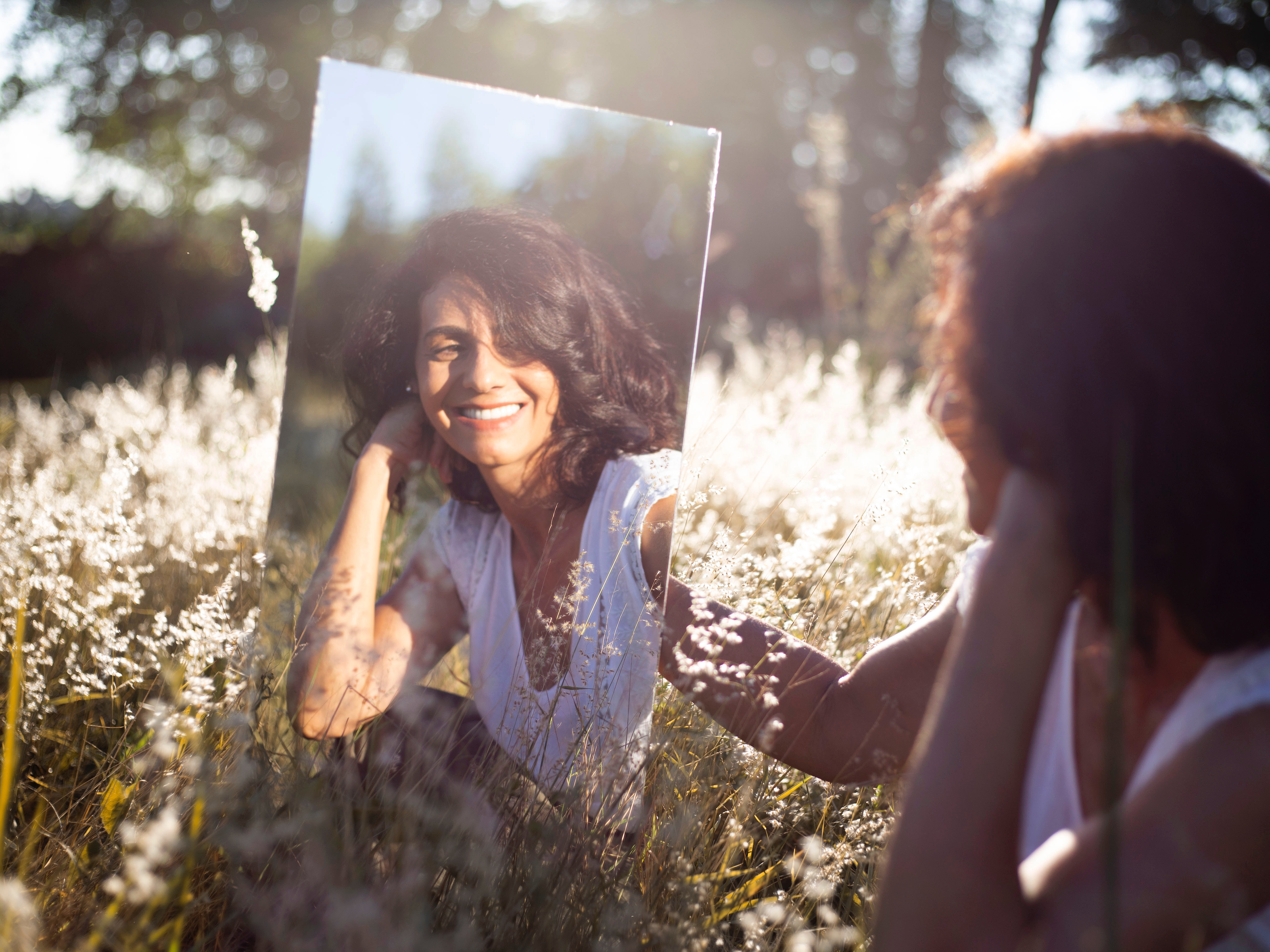 Image of a woman outdoors, smiling into a mirror whilst watching her reflection, depicting someone who is positively reflecting.