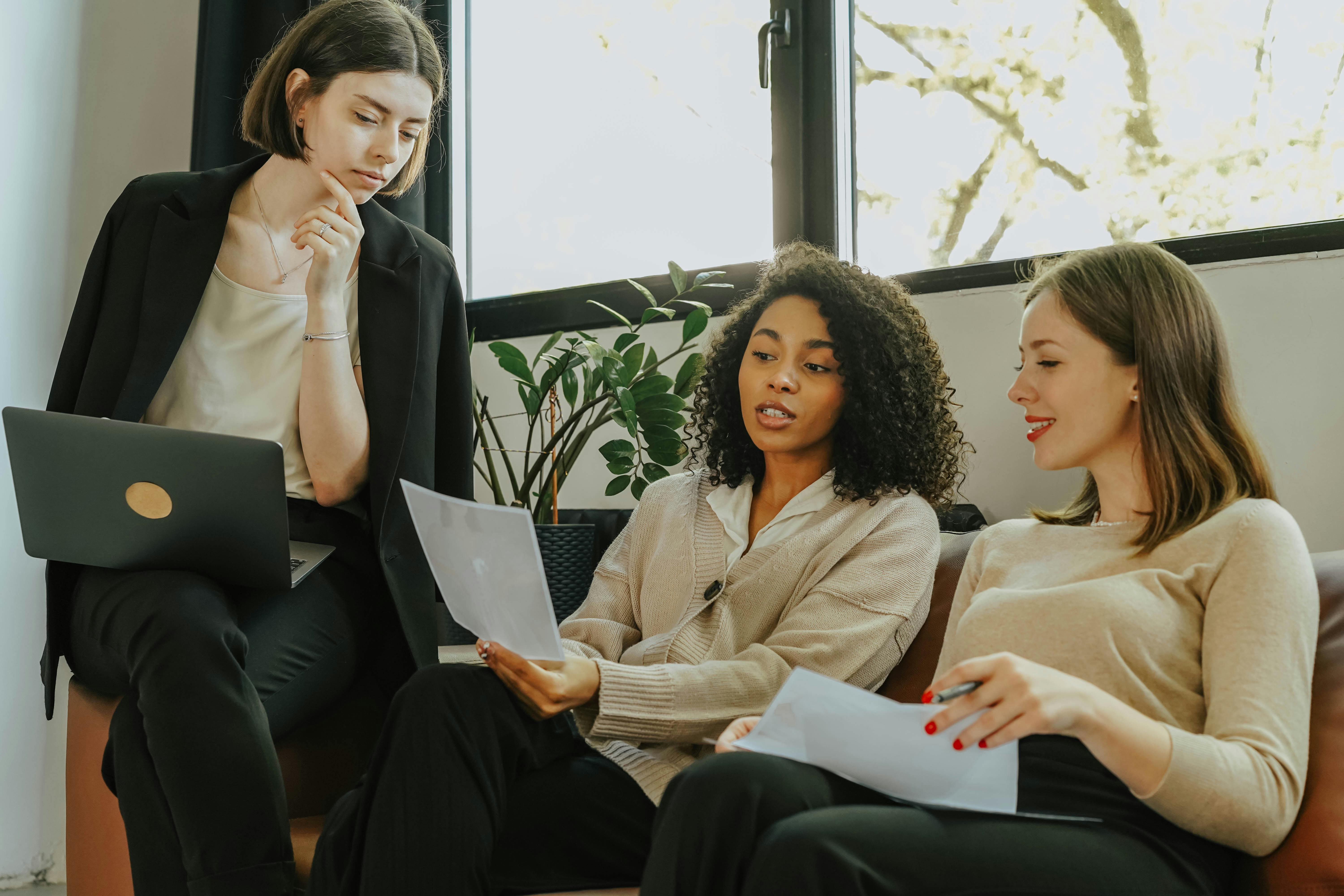 Three women sitting on a couch, each using a laptop, engaged in conversation and collaboration. Three women sitting on a couch, each using a laptop, engaged in conversation and collaboration.