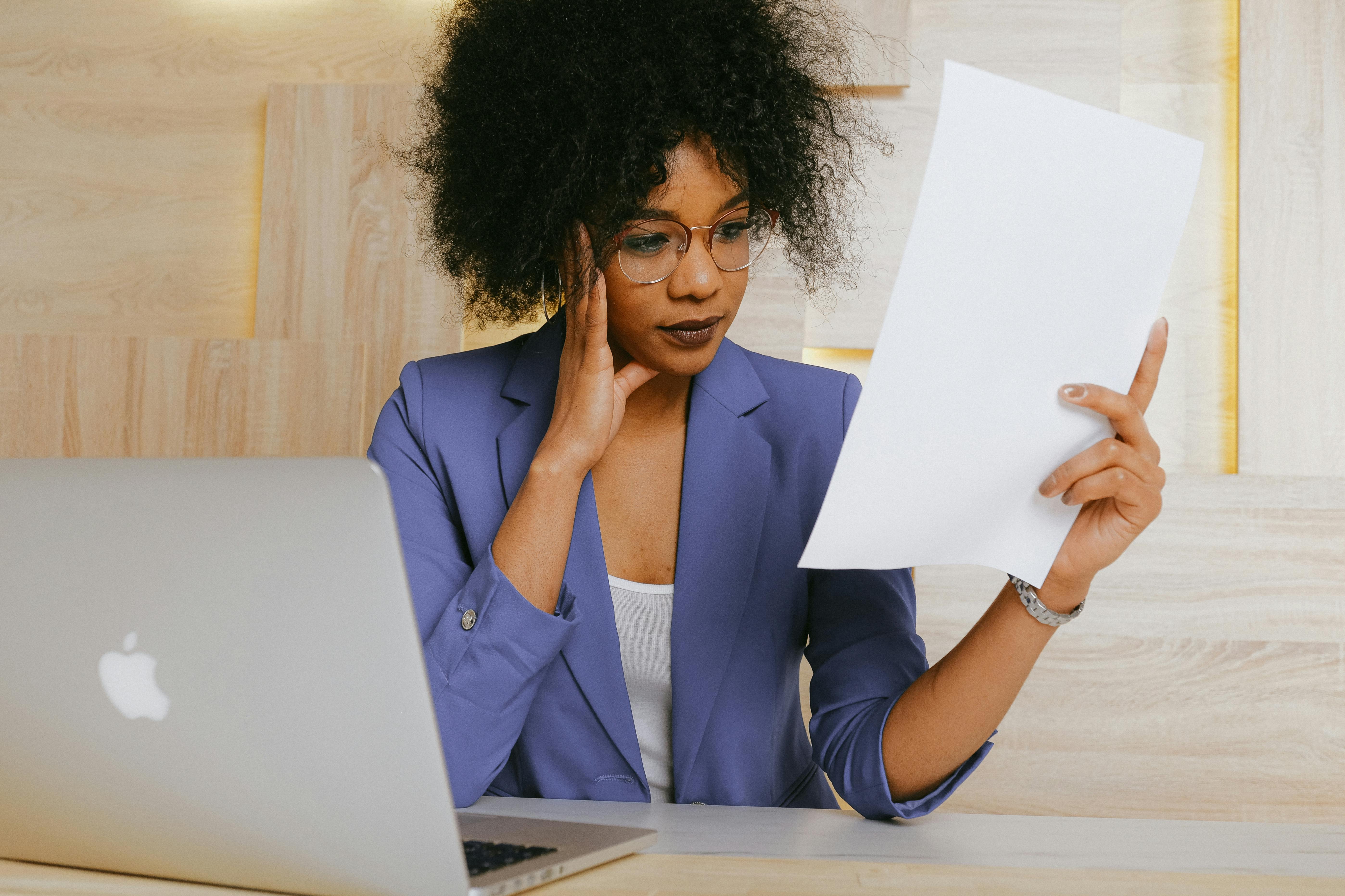 A woman in a blue blazer and glasses is holding a piece of paper, appearing engaged in reading or reviewing it. A woman in a blue blazer and glasses is holding a piece of paper, appearing engaged in reading or reviewing it.