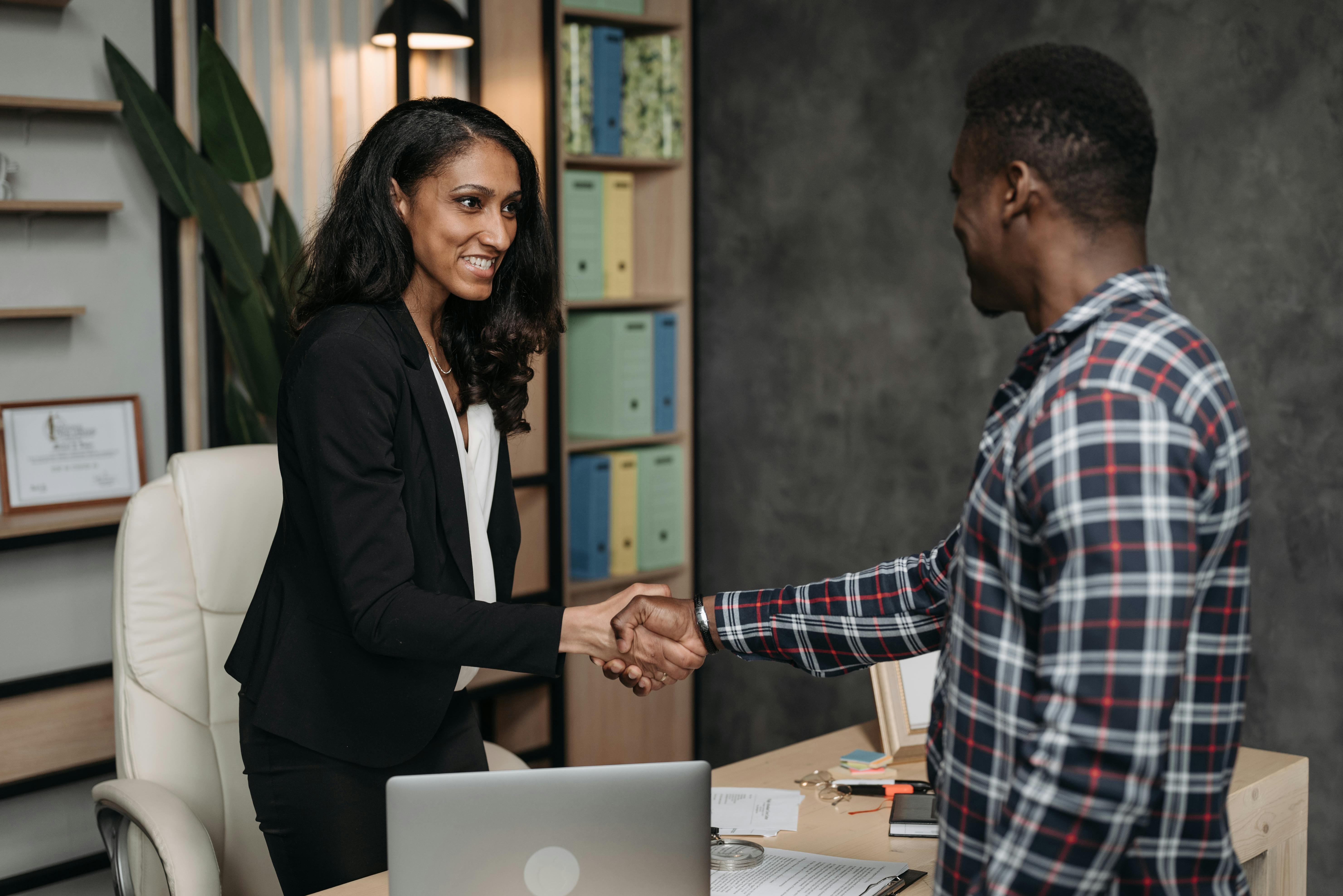 A man and woman engaged in a handshake, standing in front of a desk in a professional environment. A man and woman engaged in a handshake, standing in front of a desk in a professional environment.