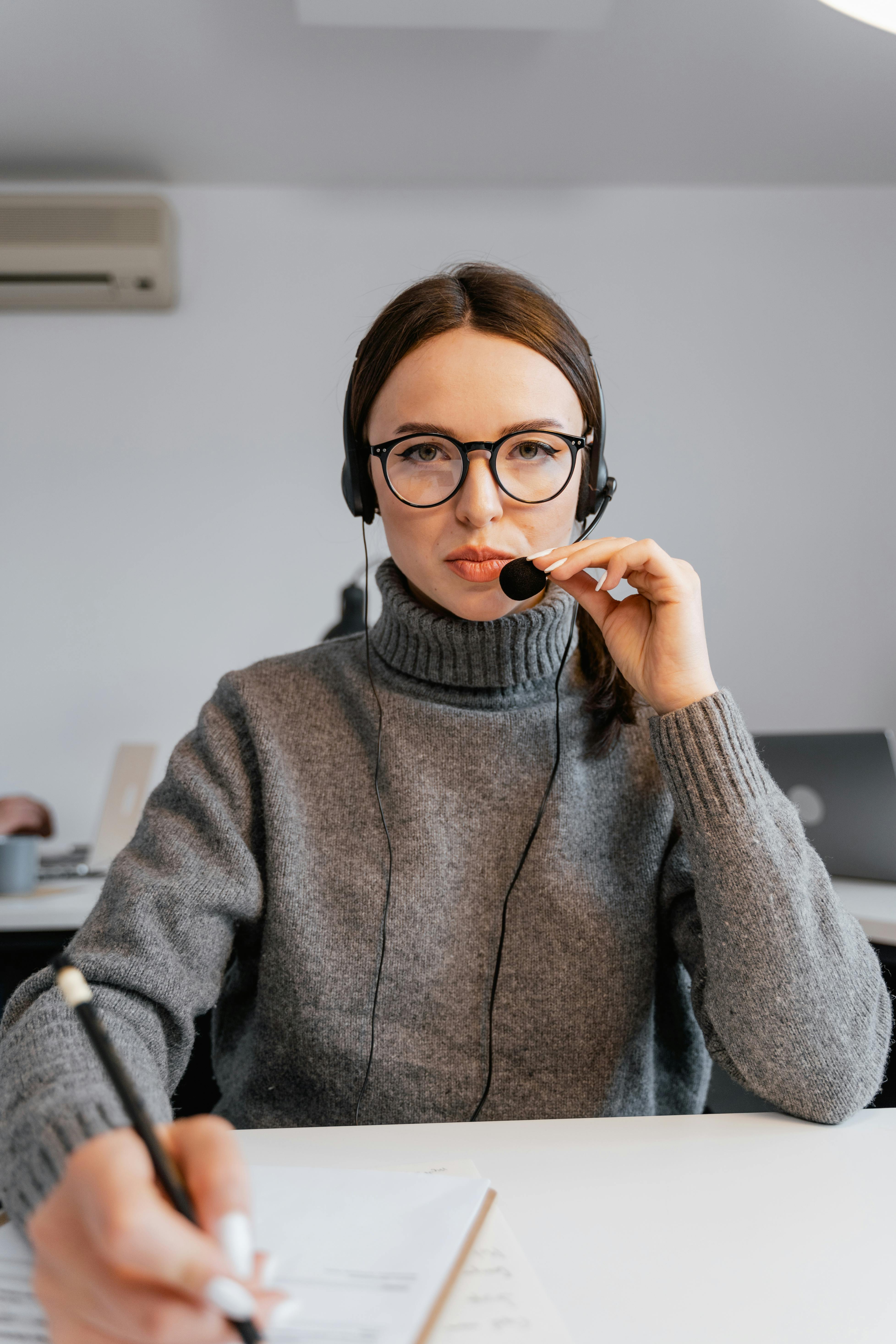 A woman in glasses and a sweater sits at a desk, writing in a notebook with a pen. A woman in glasses and a sweater sits at a desk, writing in a notebook with a pen.