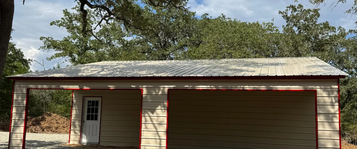 Exterior view of a beige metal garage with red trim surrounded by trees. Exterior view of a beige metal garage with red trim surrounded by trees.