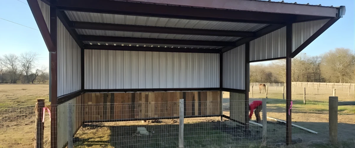Small metal barn being built on a fenced property with partial wall panels installed.