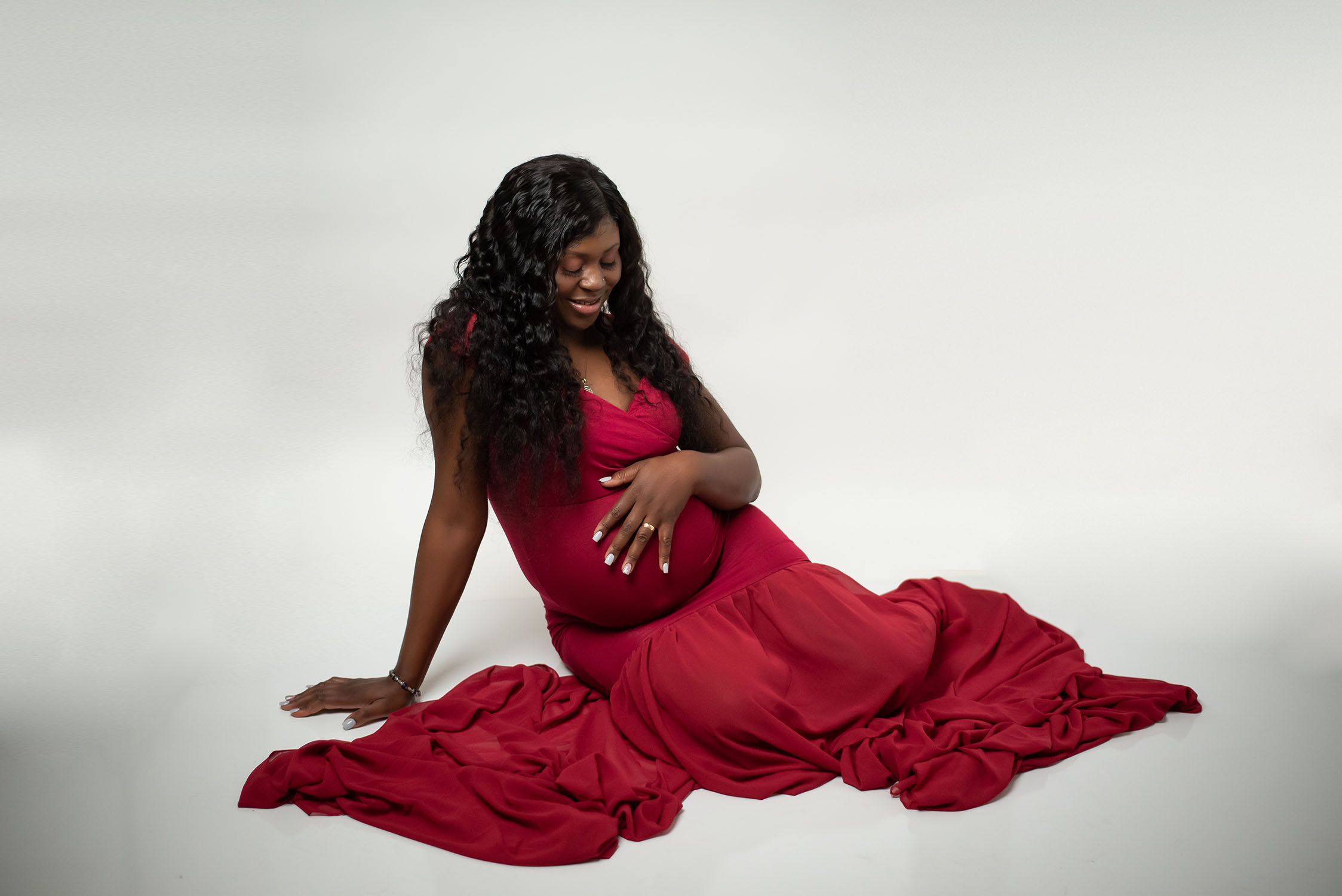 A pregnant woman with long, curly black hair sits on the floor wearing a flowing red gown. She smiles while cradling her baby bump, against a plain white backdrop. Photo taken by Leslee Shaw Photography in Sweetwater, TX.