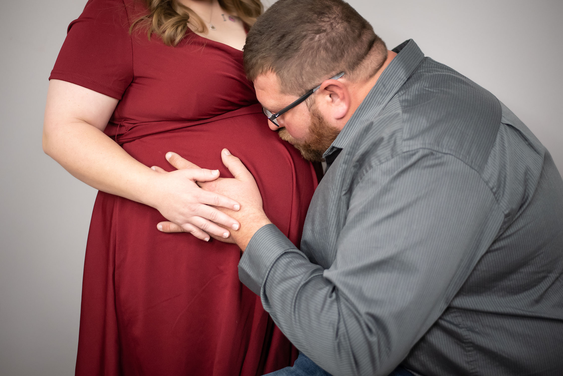 A man in a gray shirt kneels and kisses the belly of a pregnant woman wearing a red dress. Both have their hands gently placed on her baby bump, showing a moment of affection.