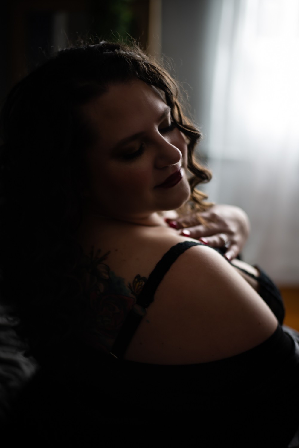 Close-up boudoir portrait of a curly-haired woman wearing black lingerie, softly smiling and showing her shoulder tattoo, photographed by Leslee Shaw Photography in Sweetwater, Texas.